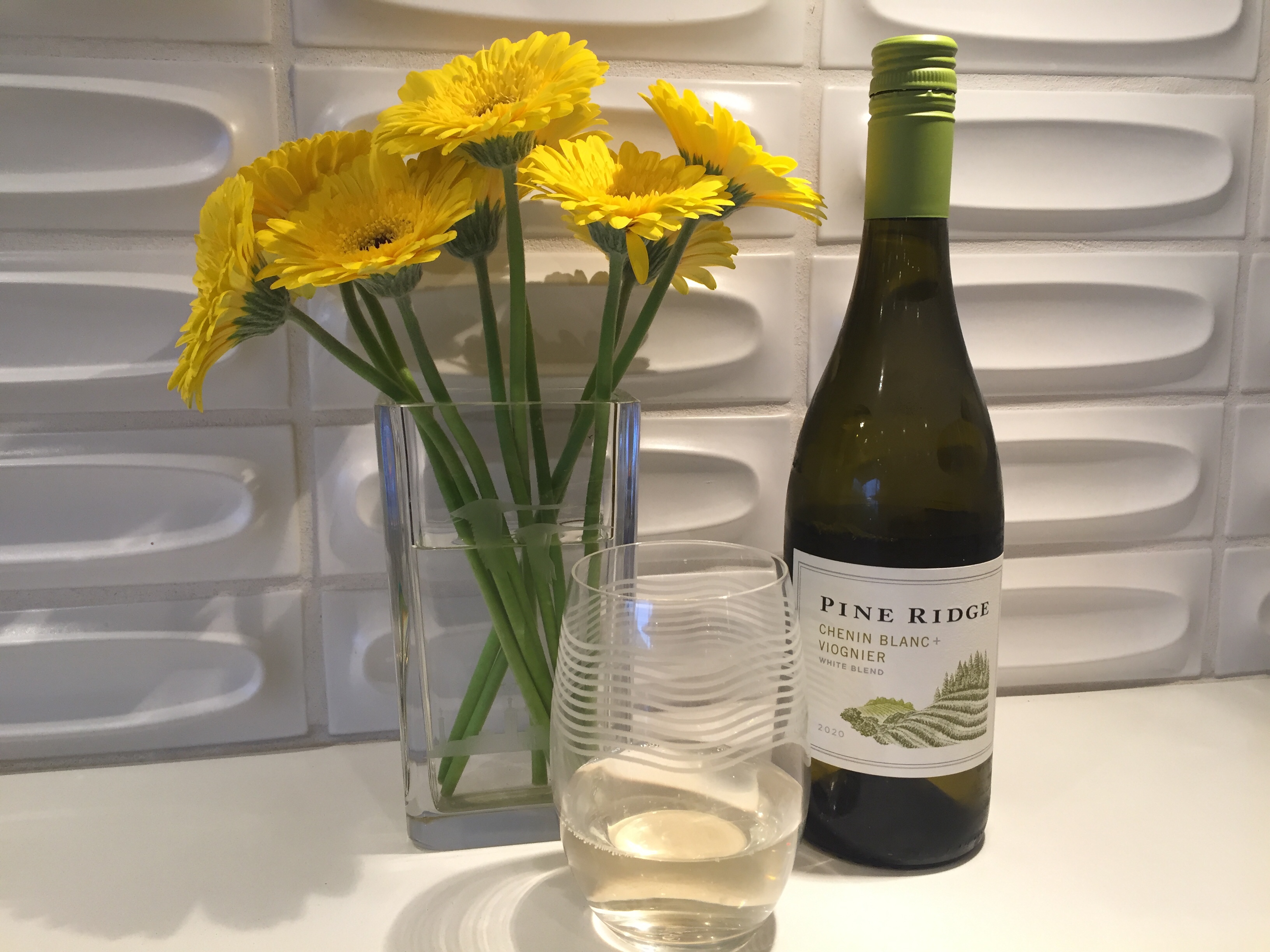Bottle and glass of Pine Ridge 2020 Chenin Blanc + Viognier - and a vase of yellow Gerberra Daisies